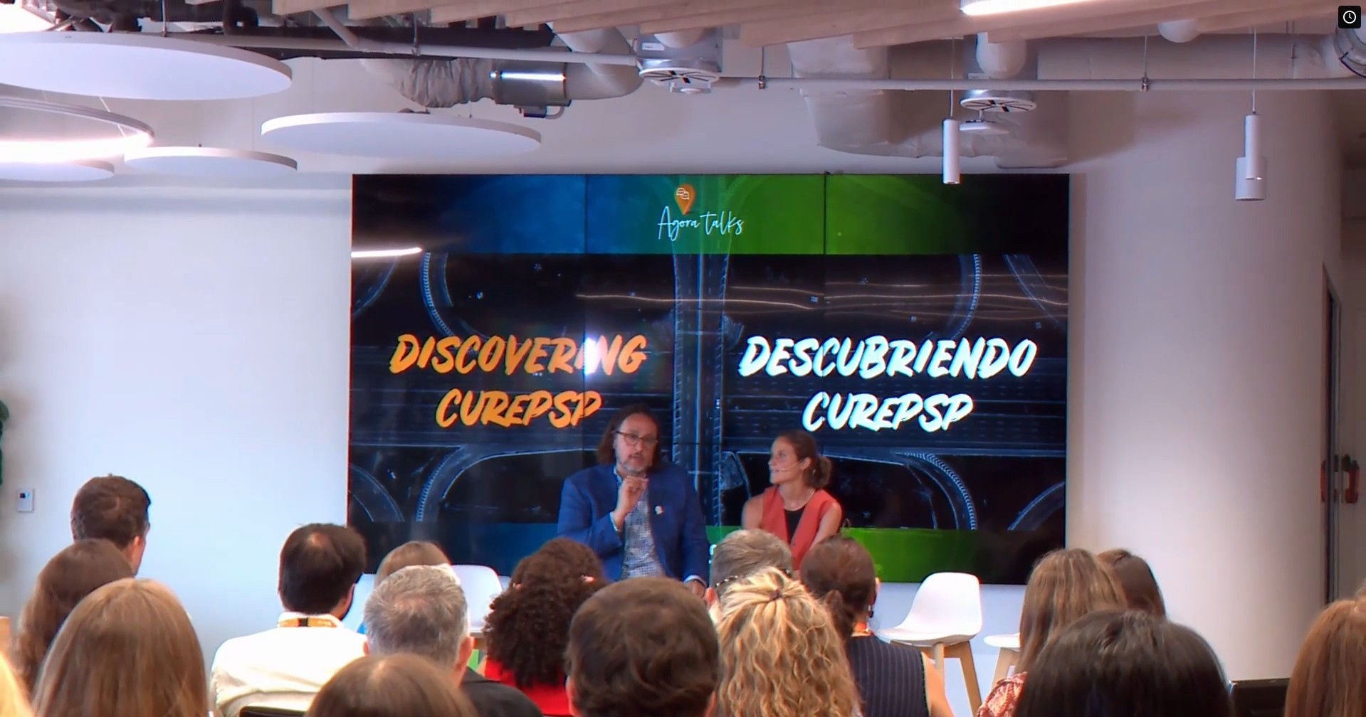 Evento corporativo de Ferrer con dos ponentes en el escenario durante una charla, frente a una audiencia, con una pantalla de fondo que muestra el mensaje “Discovering CUREP5P”.