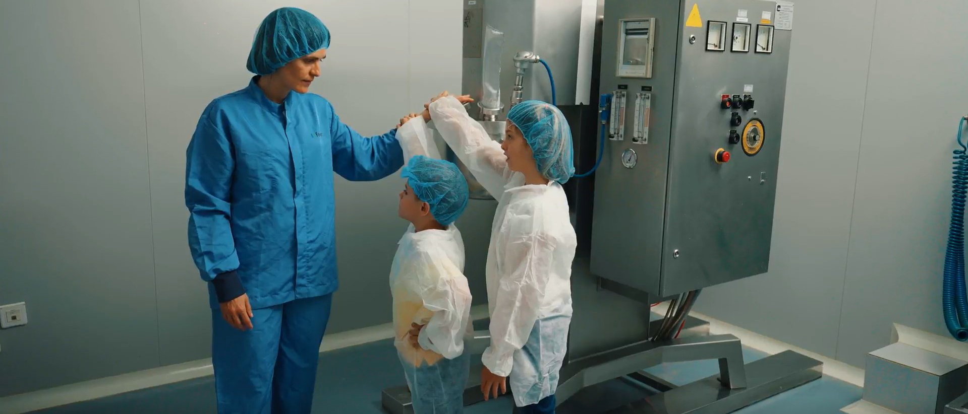 Persona de l' equip de Ferrer en una zona de producció, vestida amb uniforme i gorra de protecció, mostrant l' espai a dos infants amb bates i gorres protectors.