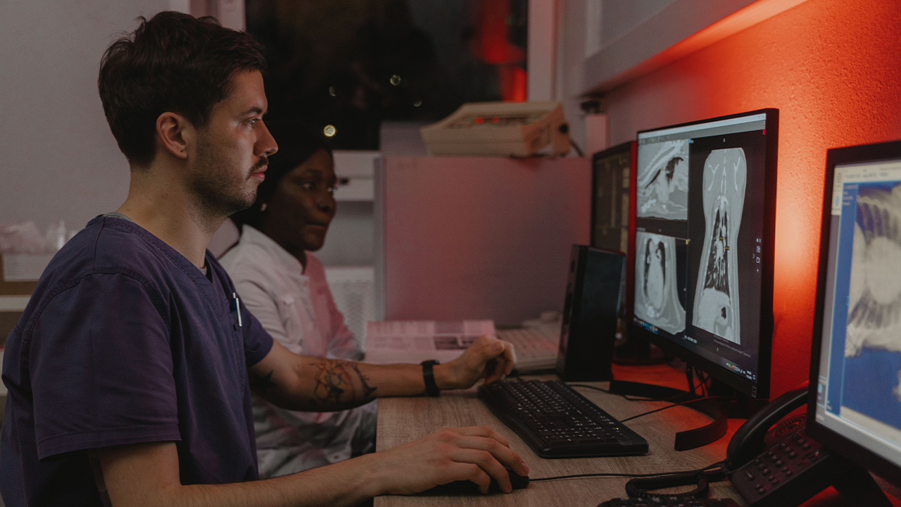 Dos professionals mèdics analitzen amb atenció imatges de tomografies i radiografies en múltiples monitors de computadora dins d'un laboratori il·luminat amb llum tènue i tons vermellosos.