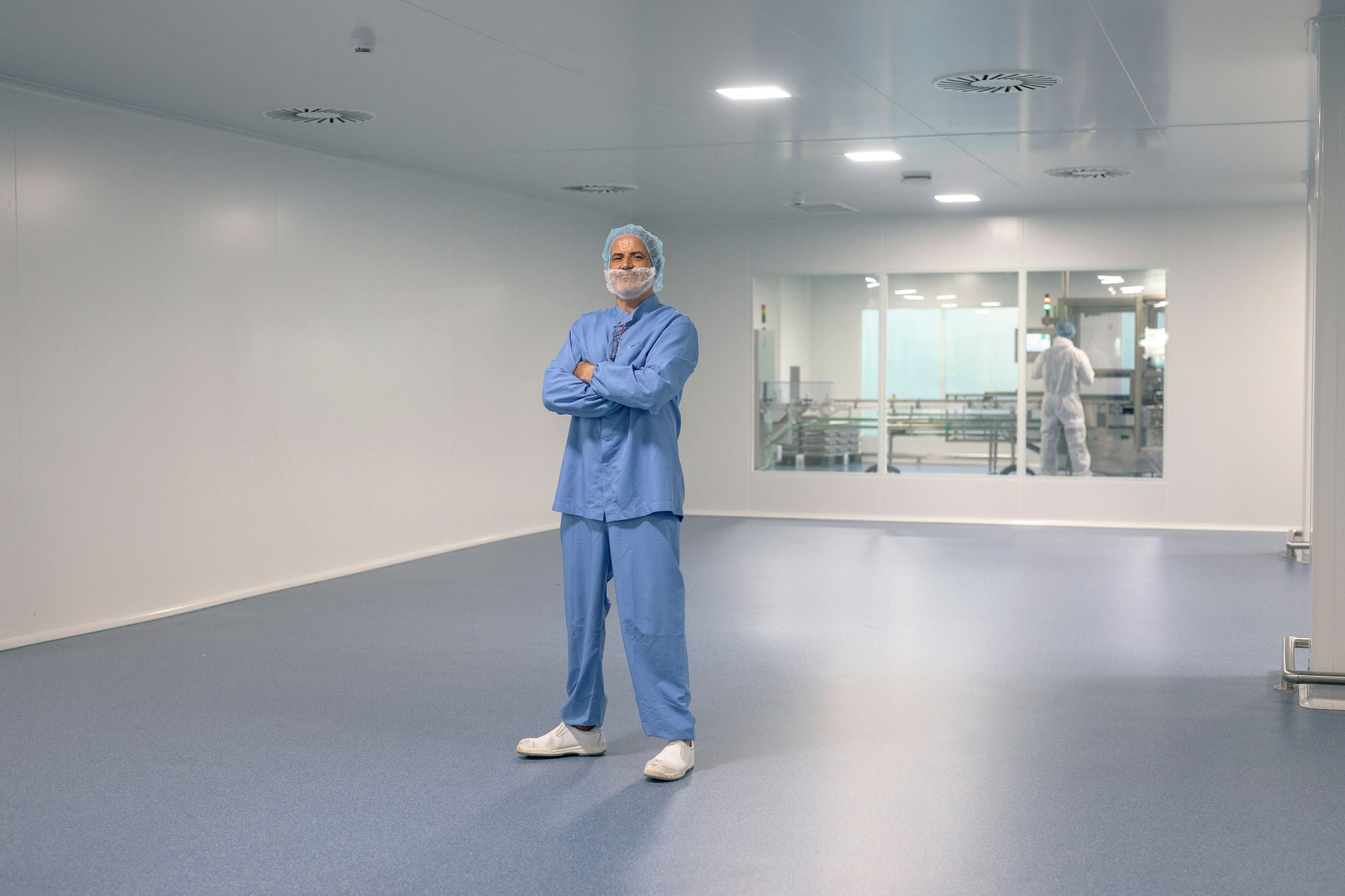 Operario con uniforme azul de protección, gorro y mascarilla posa con los brazos cruzados en una sala blanca industrial de un laboratorio o planta farmacéutica.