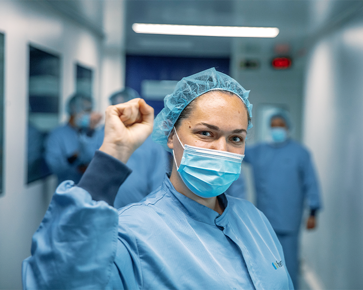 A health care professional in a uniform, mask and surgical cap smiles at the camera as she raises her fist in a hospital corridor.
