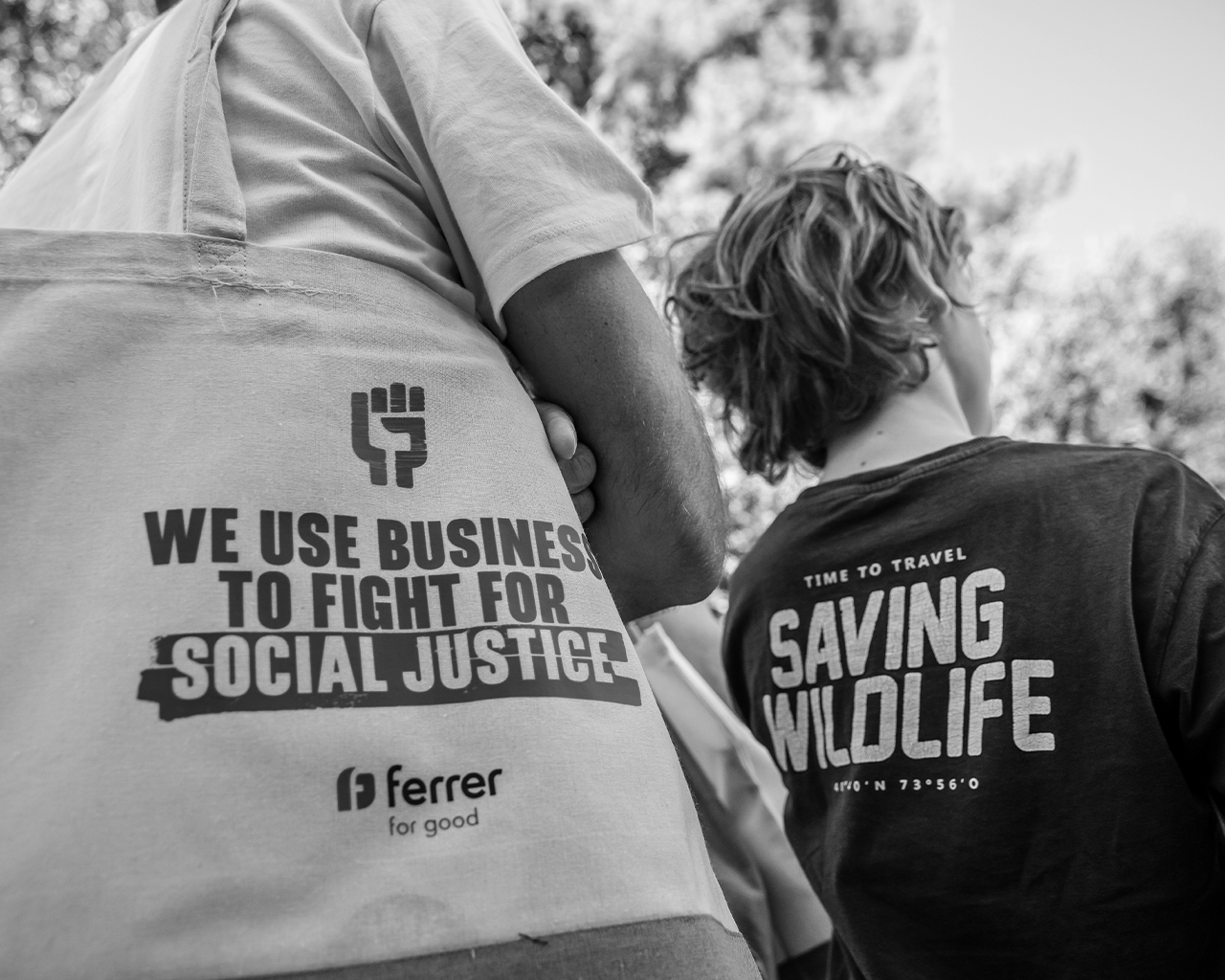Primer plano de una bolsa ecológica de tela de la empresa Ferrer for good junto a una persona con una camiseta de protección de la biodiversidad en un evento ambiental.