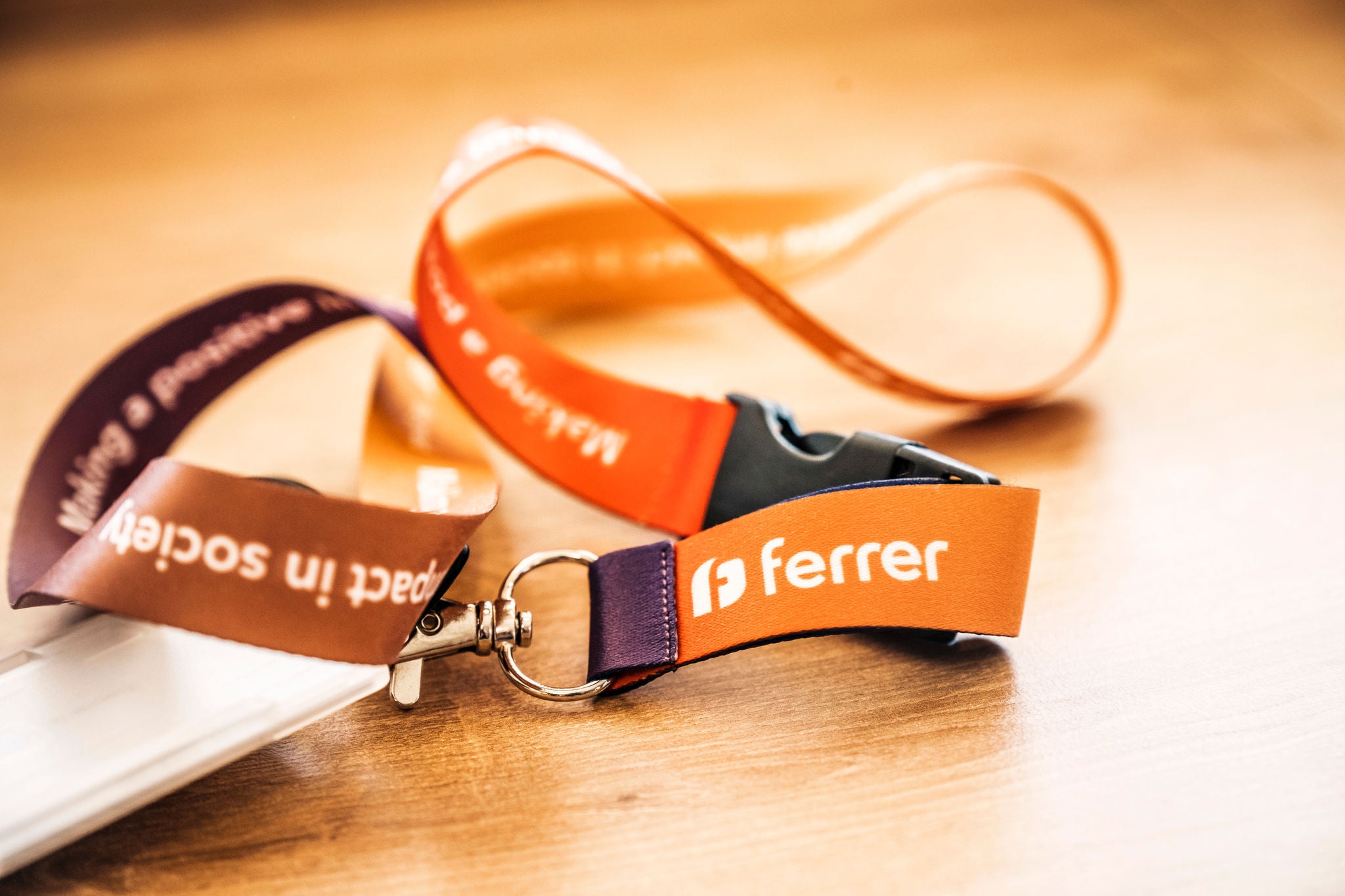 Cinta identificativa de Ferrer en tonos naranja y morado sobre una mesa de madera, con el logotipo en primer plano y un portatarjetas desenfocado a un lado.