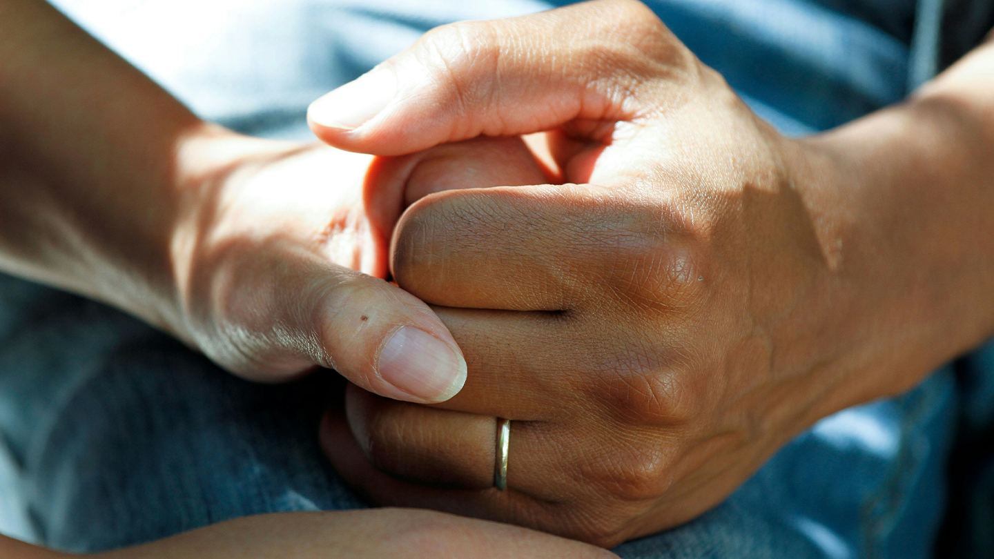 Close-up of two people tightly clasping their hands in a close gesture of support, affection or comfort.