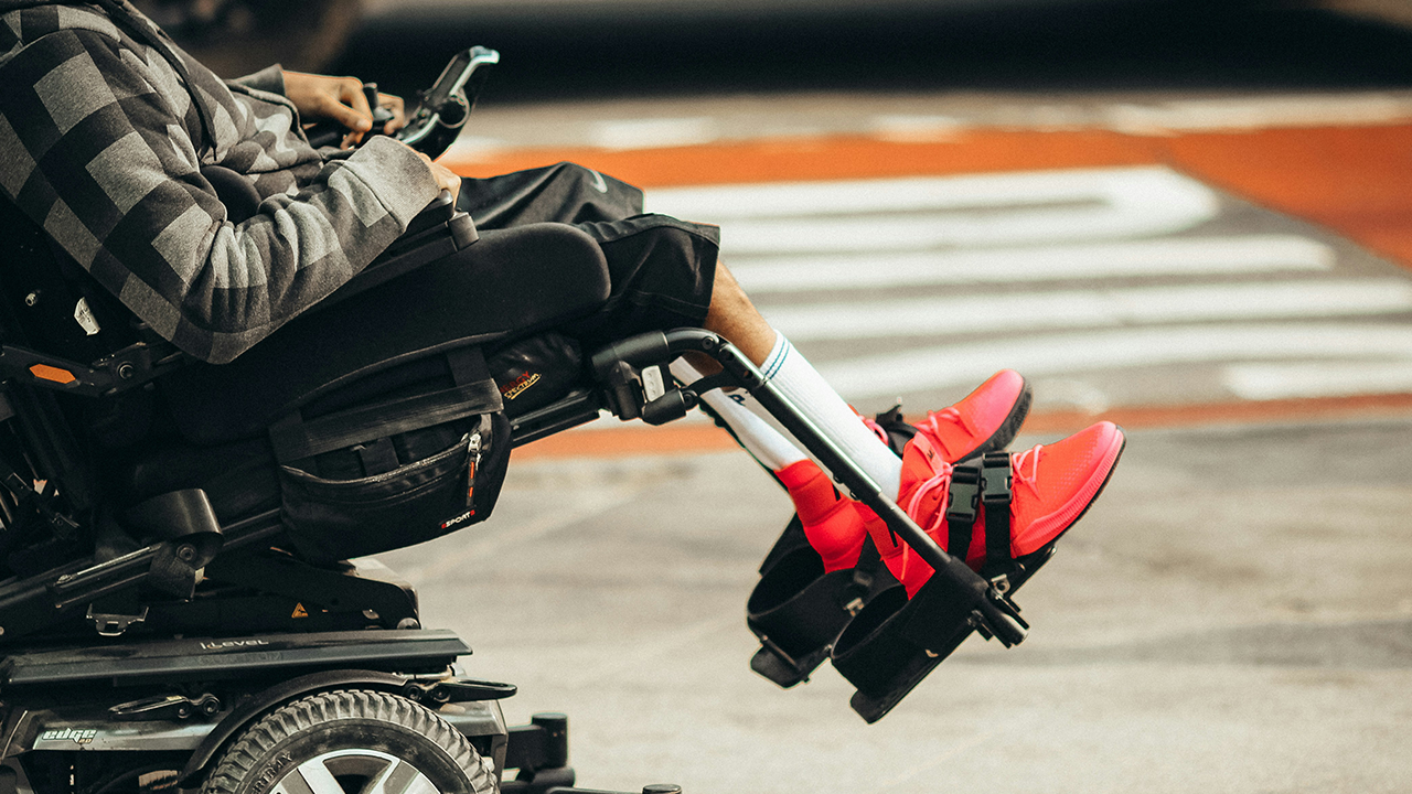 Vista de perfil y desde un ángulo bajo de una persona con zapatillas rojas brillantes sentada en una silla de ruedas motorizada en la calle.