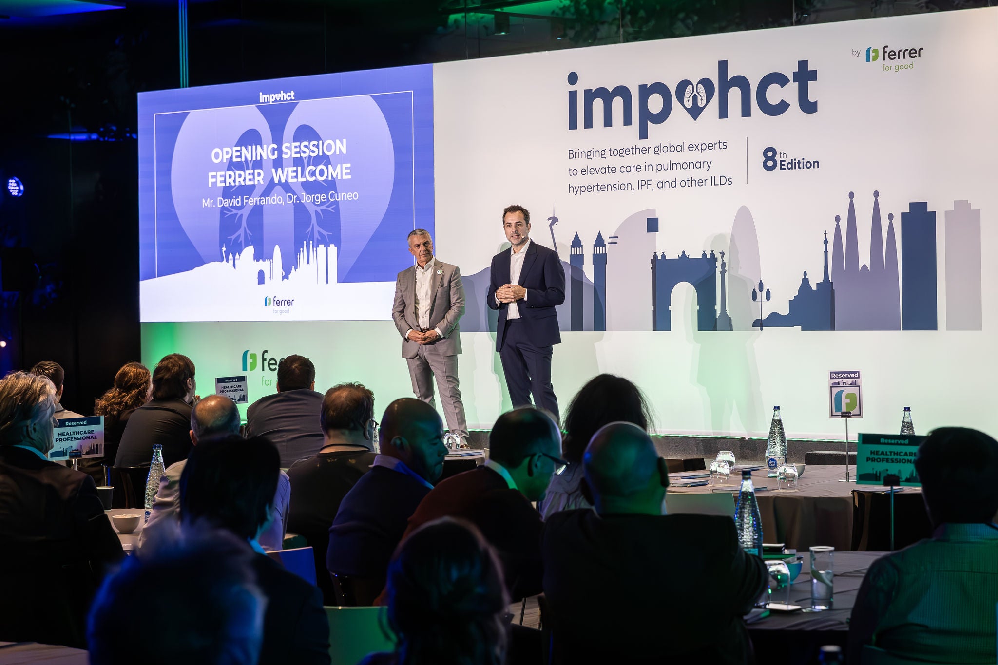 Two speakers on stage during the opening session of Ferrer's "impahct" medical event, in front of an auditorium with an audience.