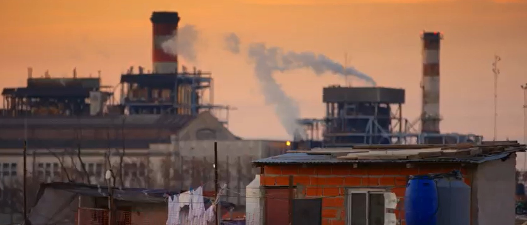 View of an industrial area at sunset with factories and chimneys emitting smoke, next to houses in the foreground.
