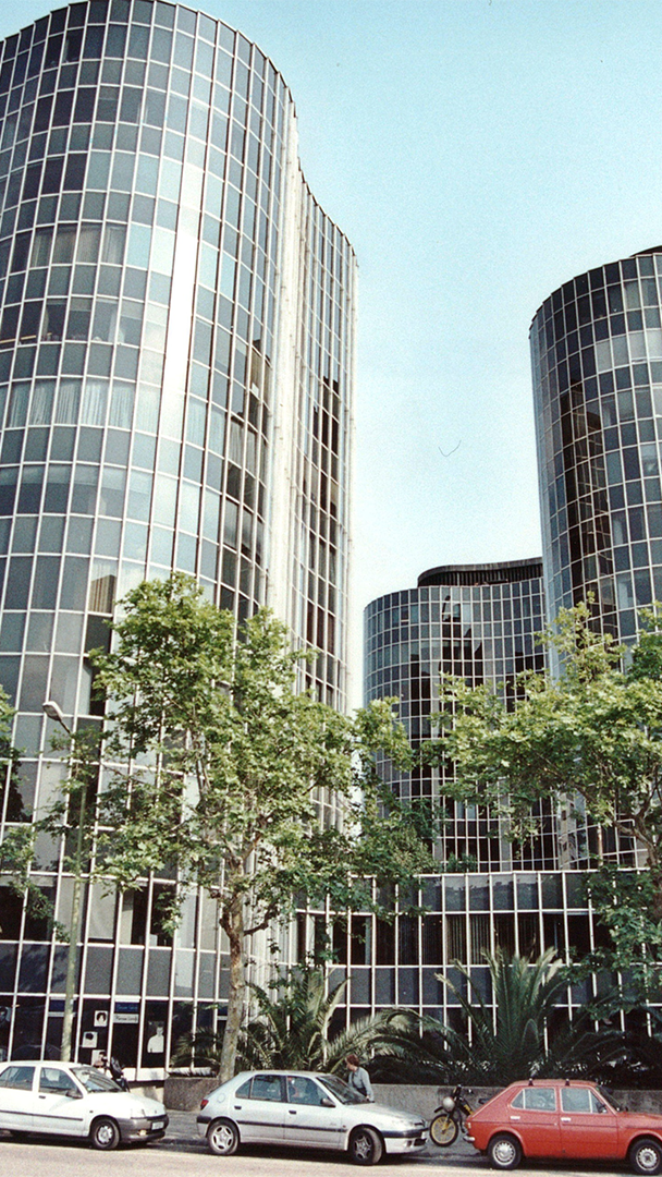 Vista en perspectiva de varios rascacielos modernos cilíndricos con fachadas acristaladas reflejantes, situados detrás de árboles y coches estacionados.