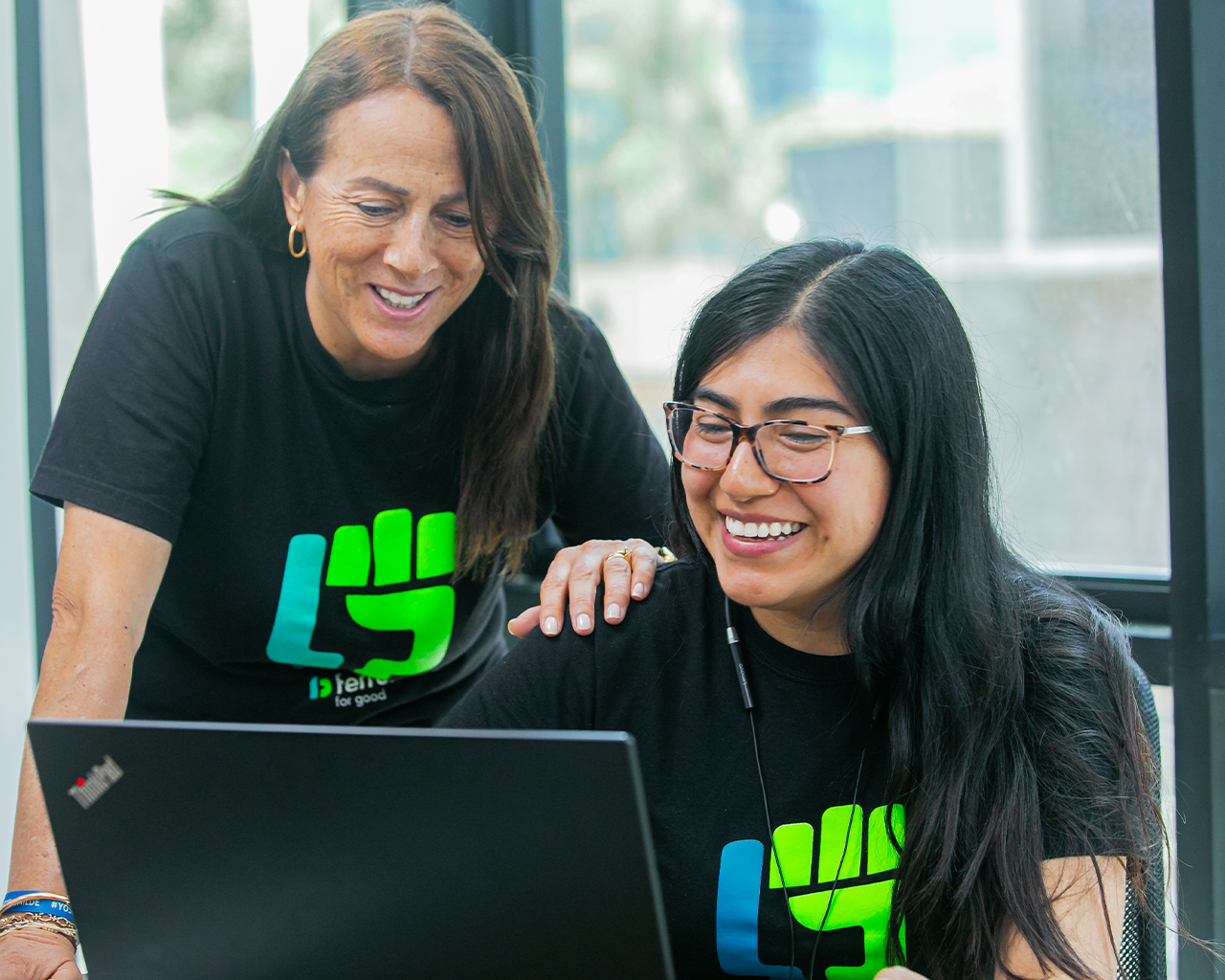 Two people from the Ferrer team work together in front of a laptop, smiling in an office environment and wearing corporate T-shirts.