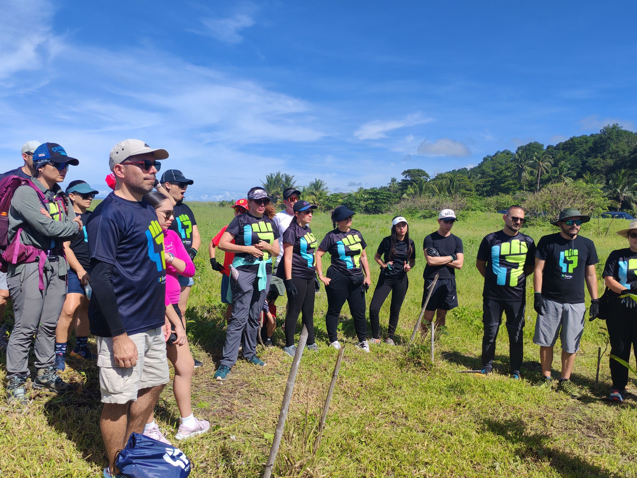 Group of people on a green field during an outdoor activity, several wearing Ferrer T-shirts, under a clear sky.