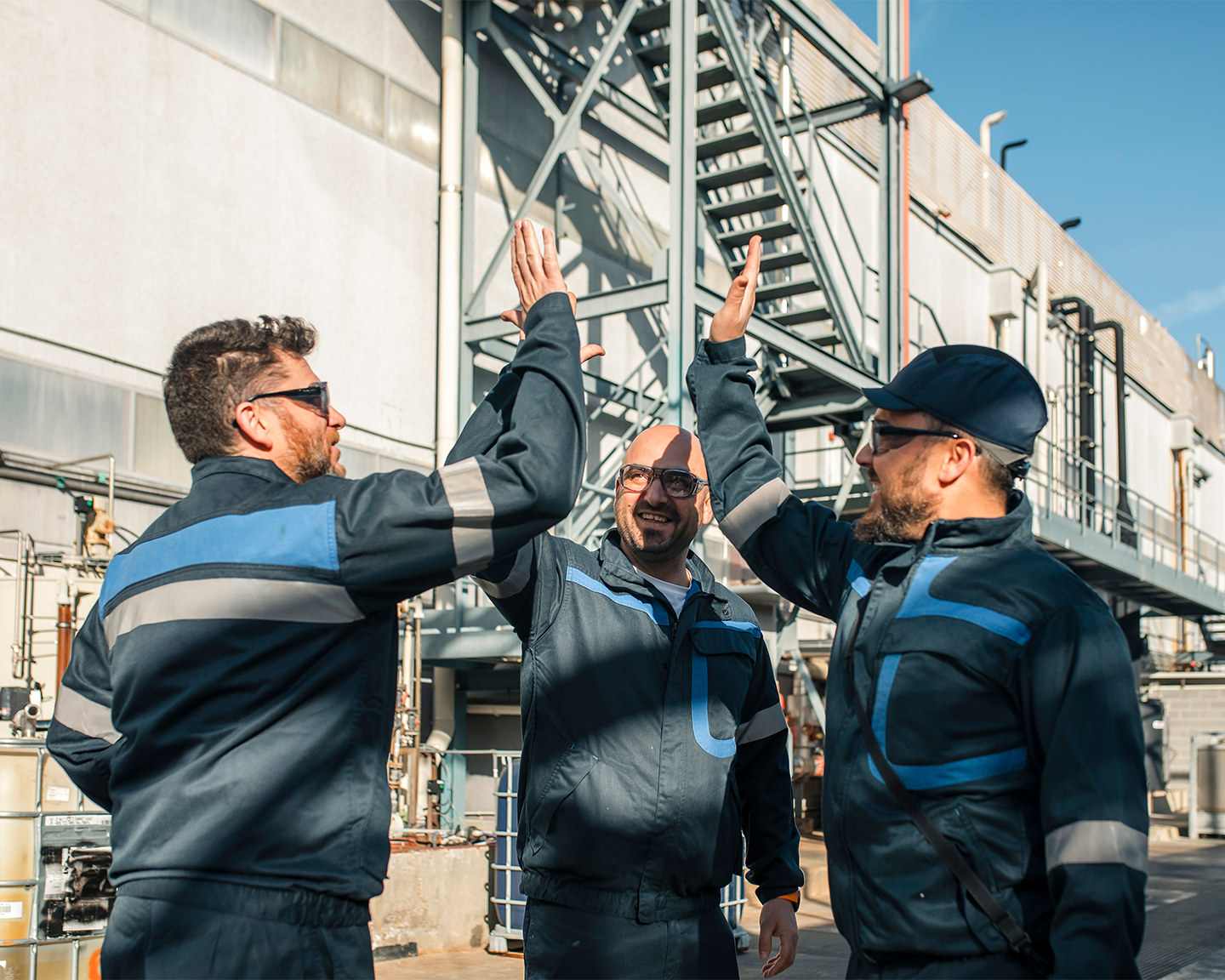 Tres operarios chocando las manos en equipo frente a las instalaciones de la fábrica industrial del Grupo Ferrer.