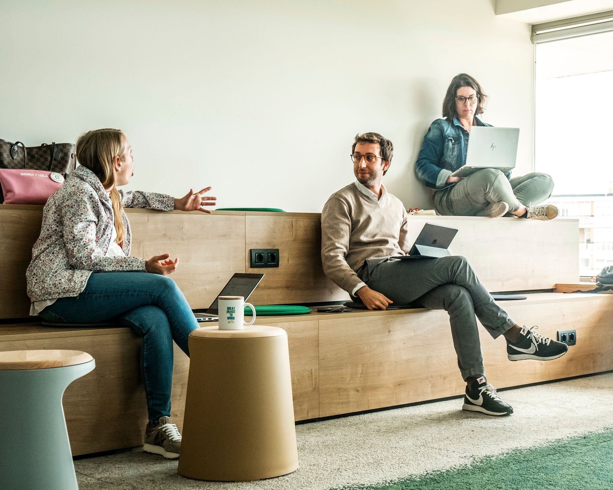 Three Ferrer professionals collaborate in an informal workspace, chatting and using laptops in an office environment.
