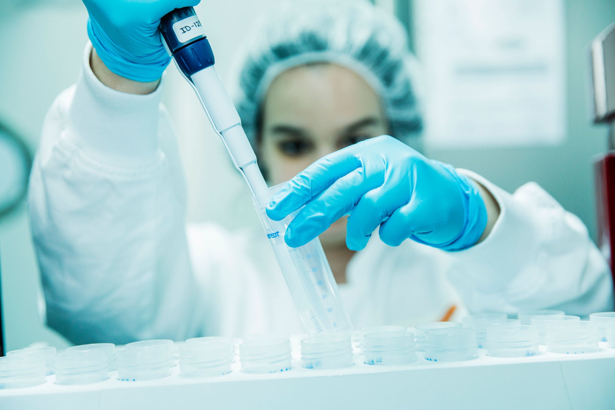Científica con guantes azules, bata y gorro protector utiliza una pipeta de precisión sobre unos viales en un laboratorio.