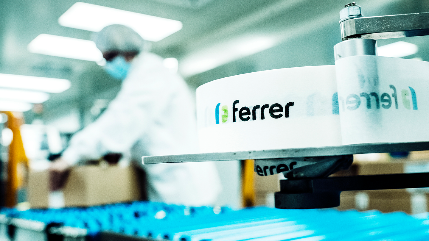 A roll of white labels with the Ferrer logo in the foreground inside a pharmaceutical packaging plant with an out-of-focus operator in the background.