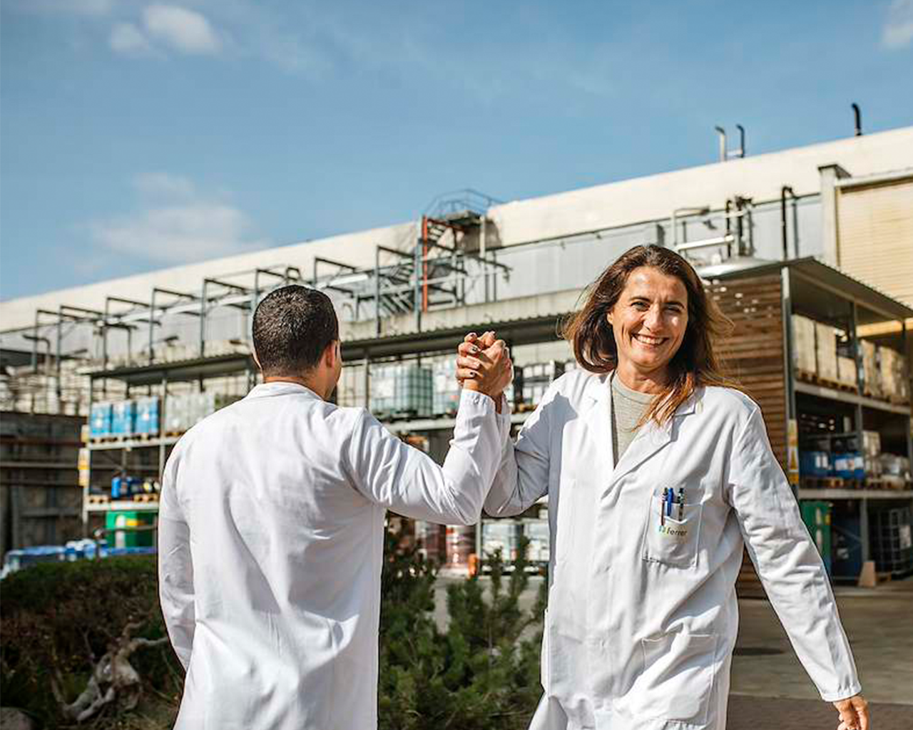 Dos profesionales con batas blancas de laboratorio se saludan con un choque de manos o pulso en el exterior de unas instalaciones industriales.