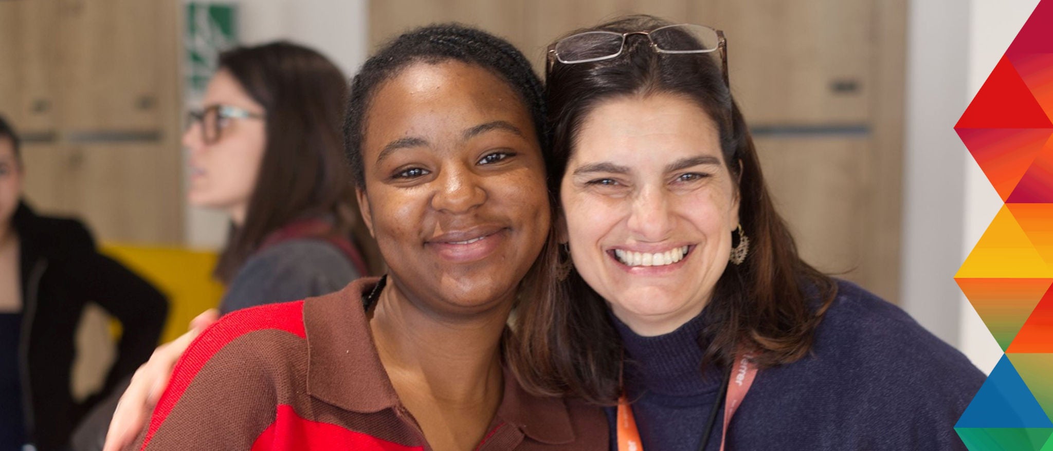 Two women smiling and embracing in an indoor setting, with a colorful geometric design on the right side of the image.
