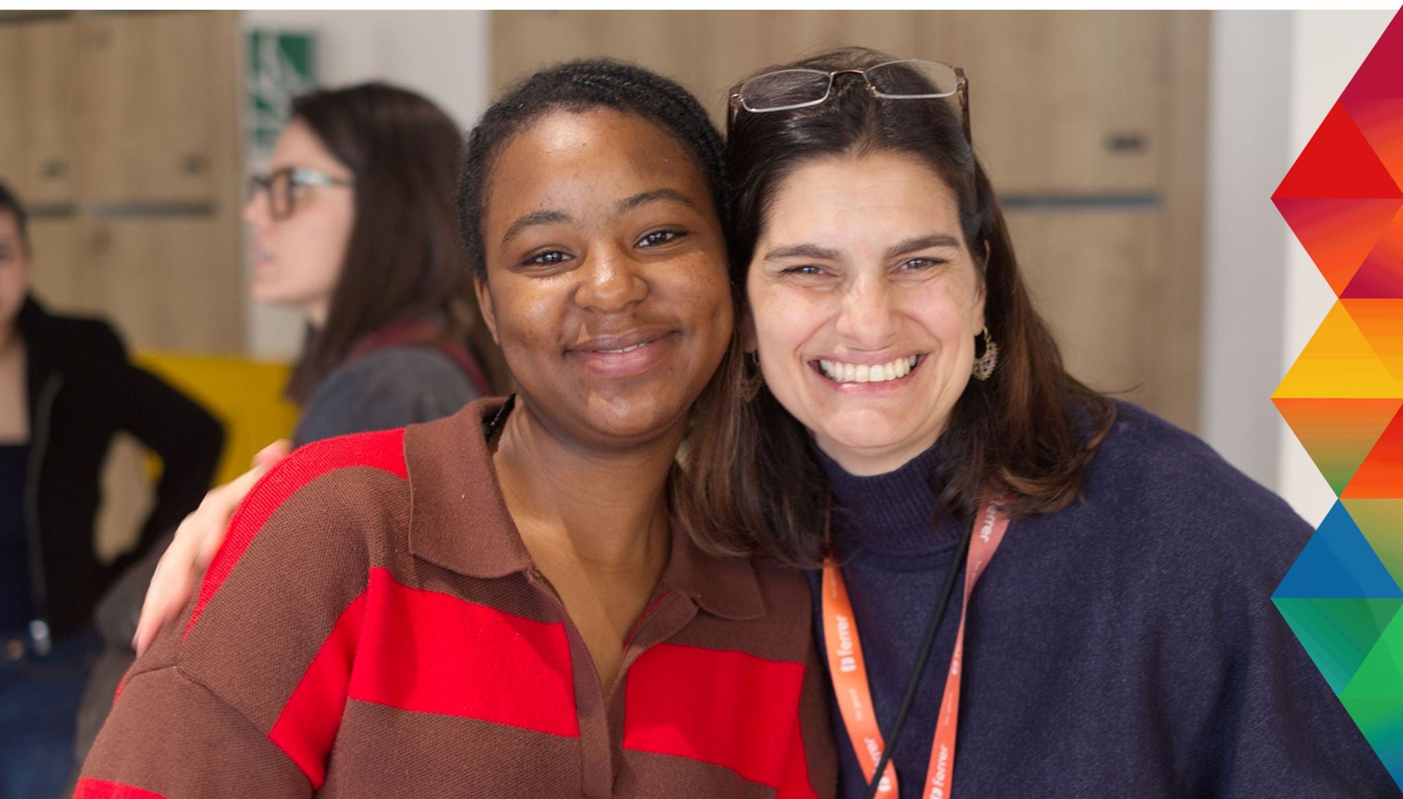 Two women smiling and embracing in an indoor setting, with a colorful geometric design on the right side of the image.