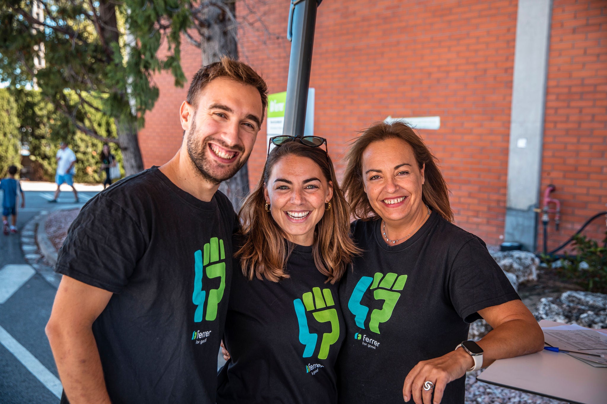 Tres voluntarios de la empresa farmacéutica Ferrer sonríen y posan juntos al aire libre, vistiendo camisetas negras con el logo verde de una iniciativa de voluntariado.