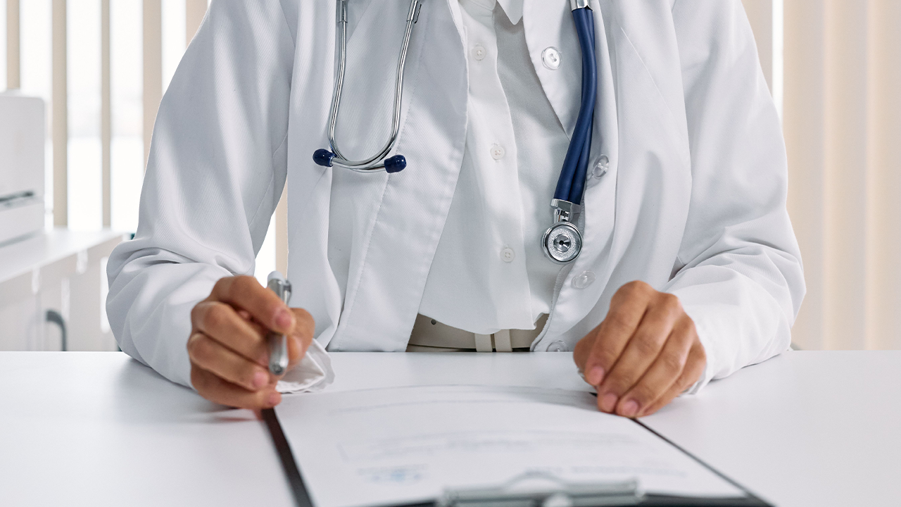 Una médica con bata blanca y estetoscopio sostiene un bolígrafo sobre un historial clínico en su escritorio.