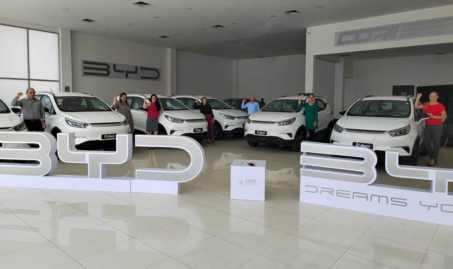 BYD car dealership with several white electric vehicles exposed and a group of people posing next to them with their fists raised.