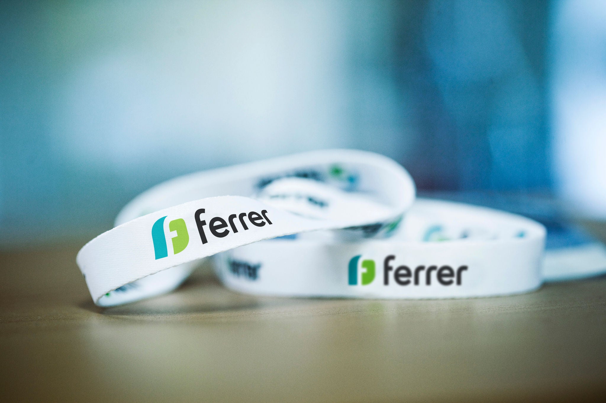 Pulseras blancas de Ferrer sobre una mesa, con el logotipo en negro, verde y azul en primer plano y el fondo desenfocado.