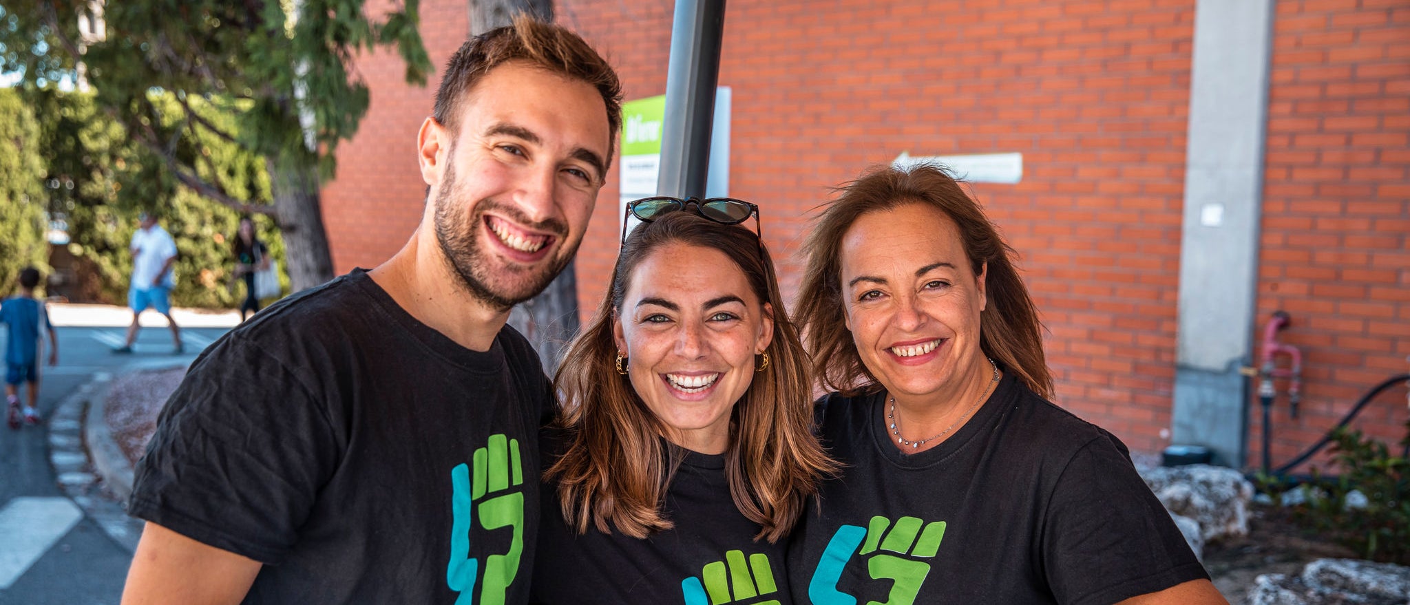 Tres voluntaris de l'empresa farmacèutica Ferrer somriuen i posen junts a l'aire lliure, vestint samarretes negres amb el logo verd d'una iniciativa de voluntariat.