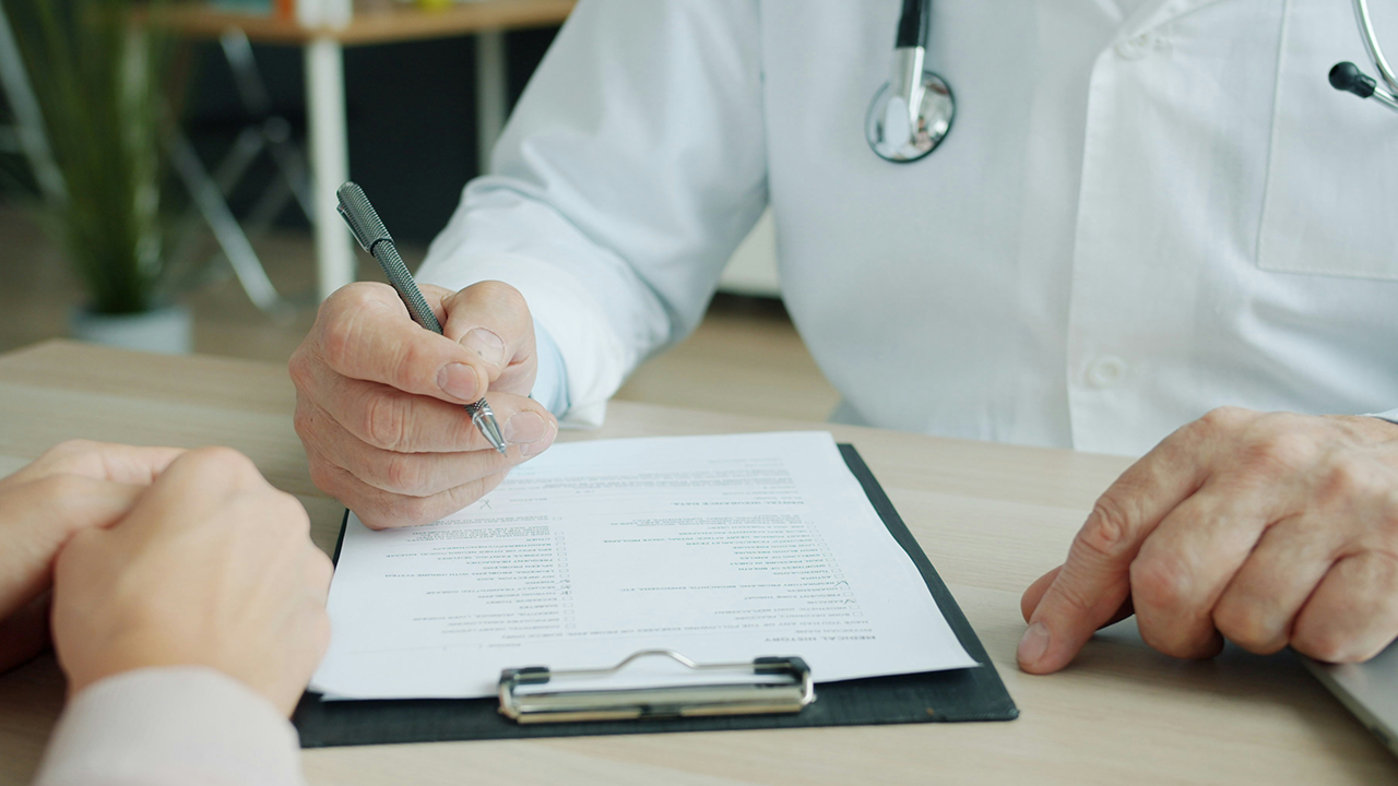 Close-up of a doctor in a white coat with a stethoscope holding a pen over a medical history on a clipboard as he attends to a patient at his desk.