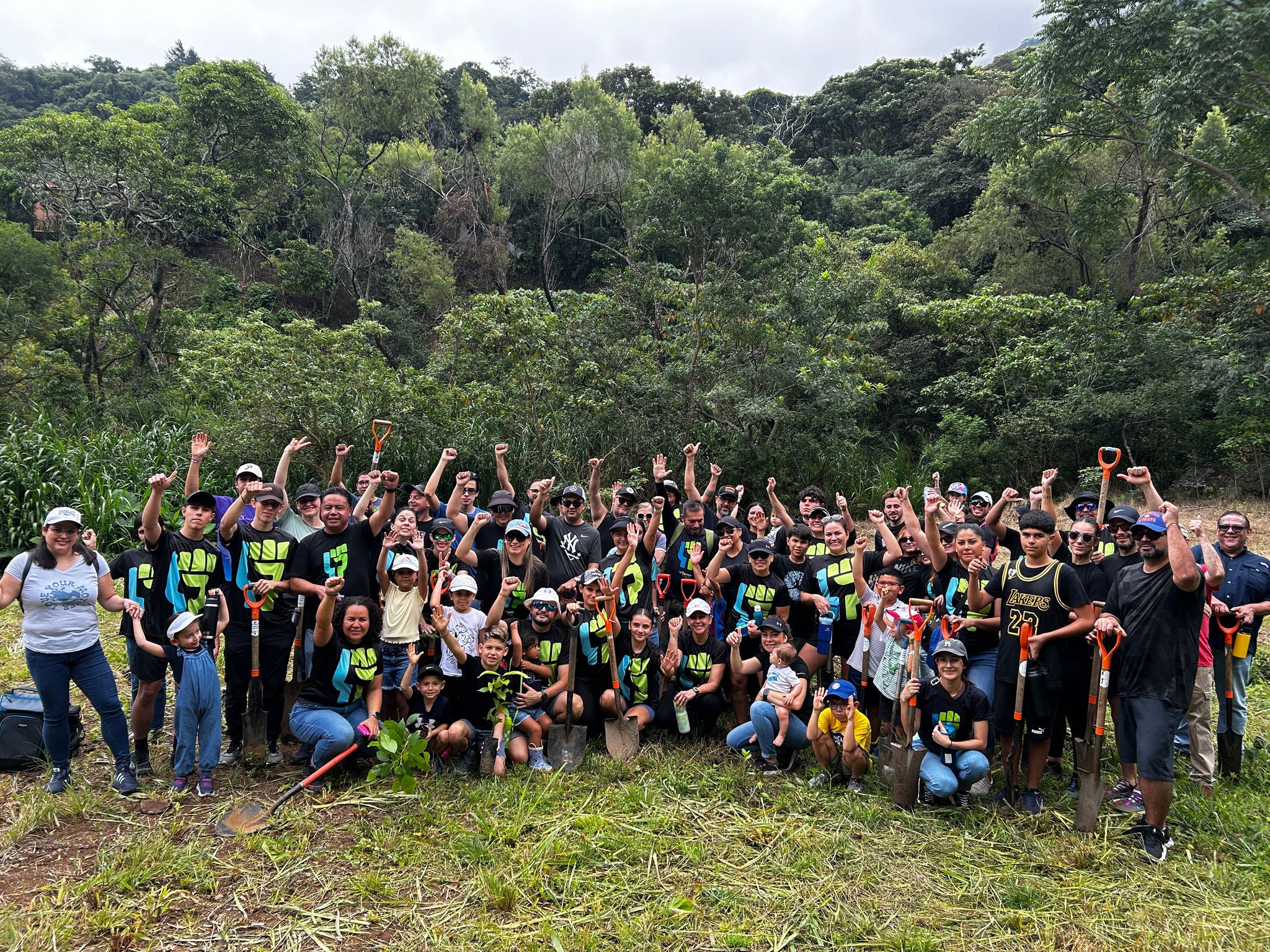 Voluntarios de Ferrer posan en grupo en una zona verde y boscosa, con palas y plantas, celebrando una jornada de reforestación o voluntariado ambiental.