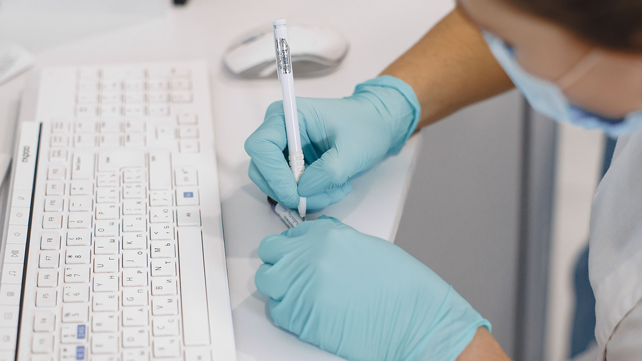 Una persona con guantes médicos azules y mascarilla escribe con un bolígrafo sobre un escritorio junto a un teclado blanco.