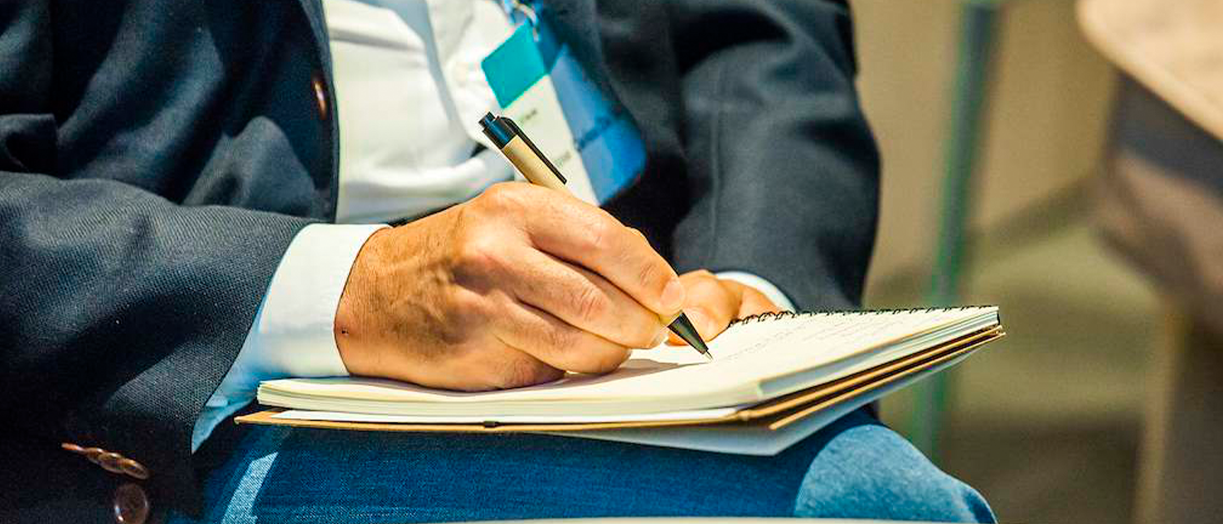 Close-up of a person in a dark jacket taking notes with a pen in a notebook resting on his lap during a corporate event or conference.