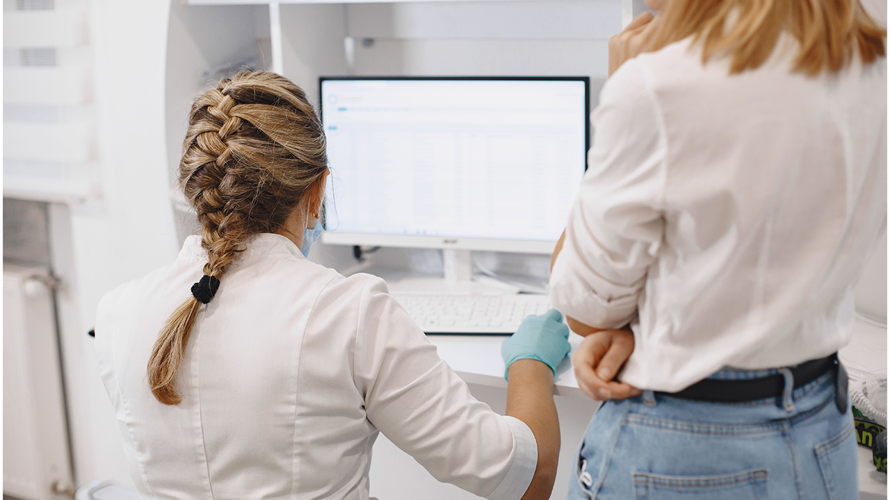 Vista posterior d'una professional mèdica amb els cabells trenats treballant davant una computadora al costat d'una col·lega en un consultori o laboratori.