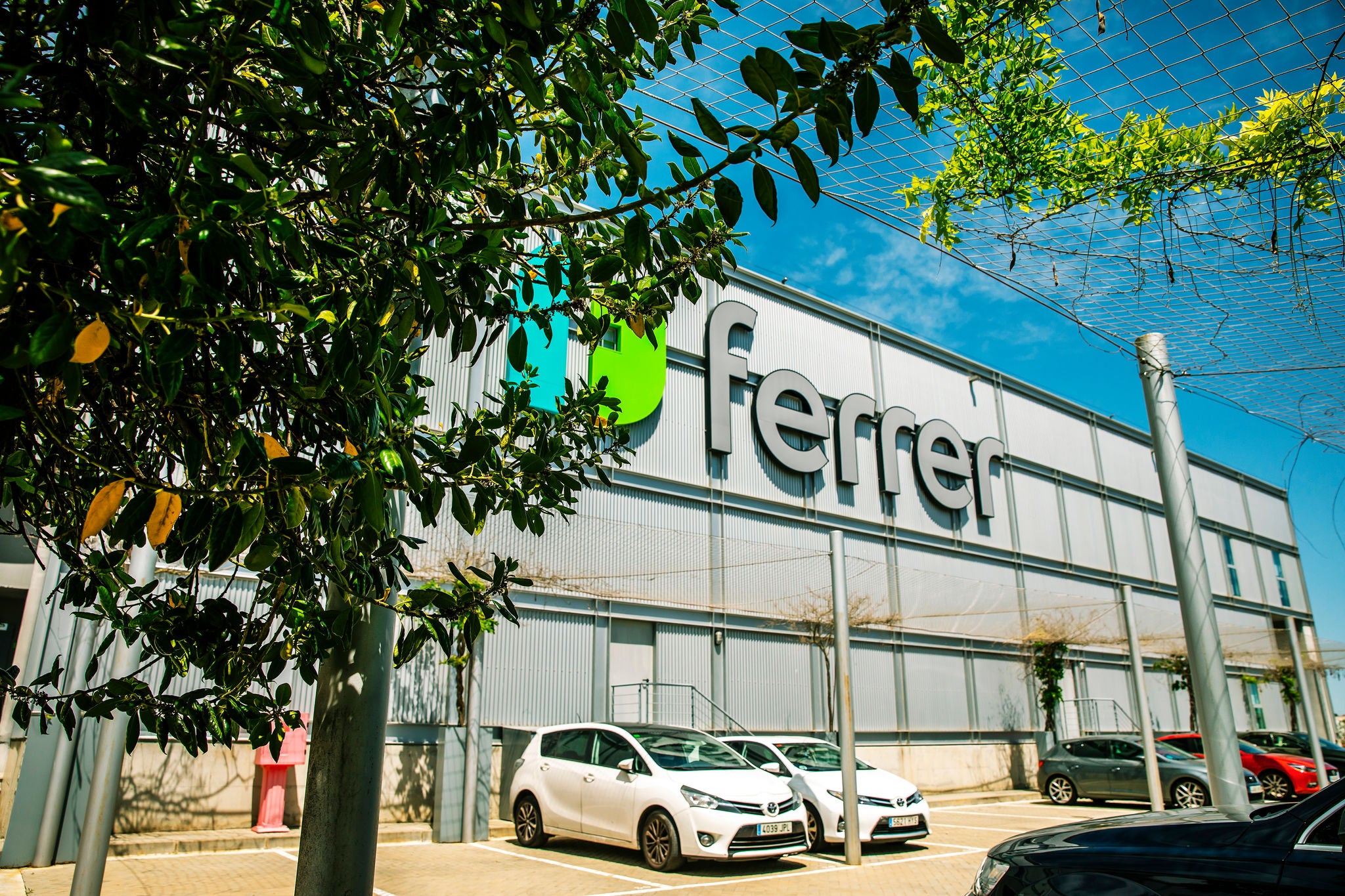 Façade of the Ferrer logistics centre with the company's logo, parked vehicles and surrounding green areas.