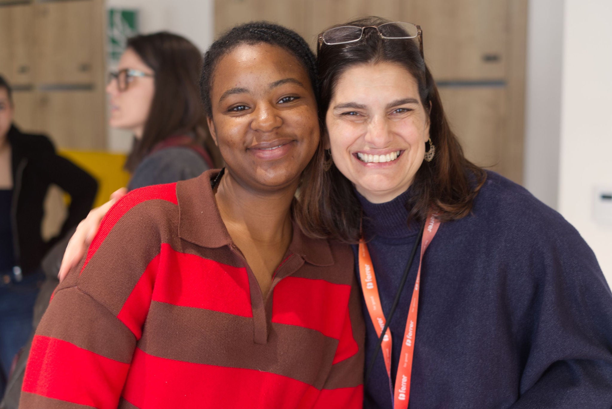 Two smiling women pose embracing in front of the camera in an indoor space, one wearing a red and brown striped polo shirt and the other a navy blue sweater with glasses over her head.