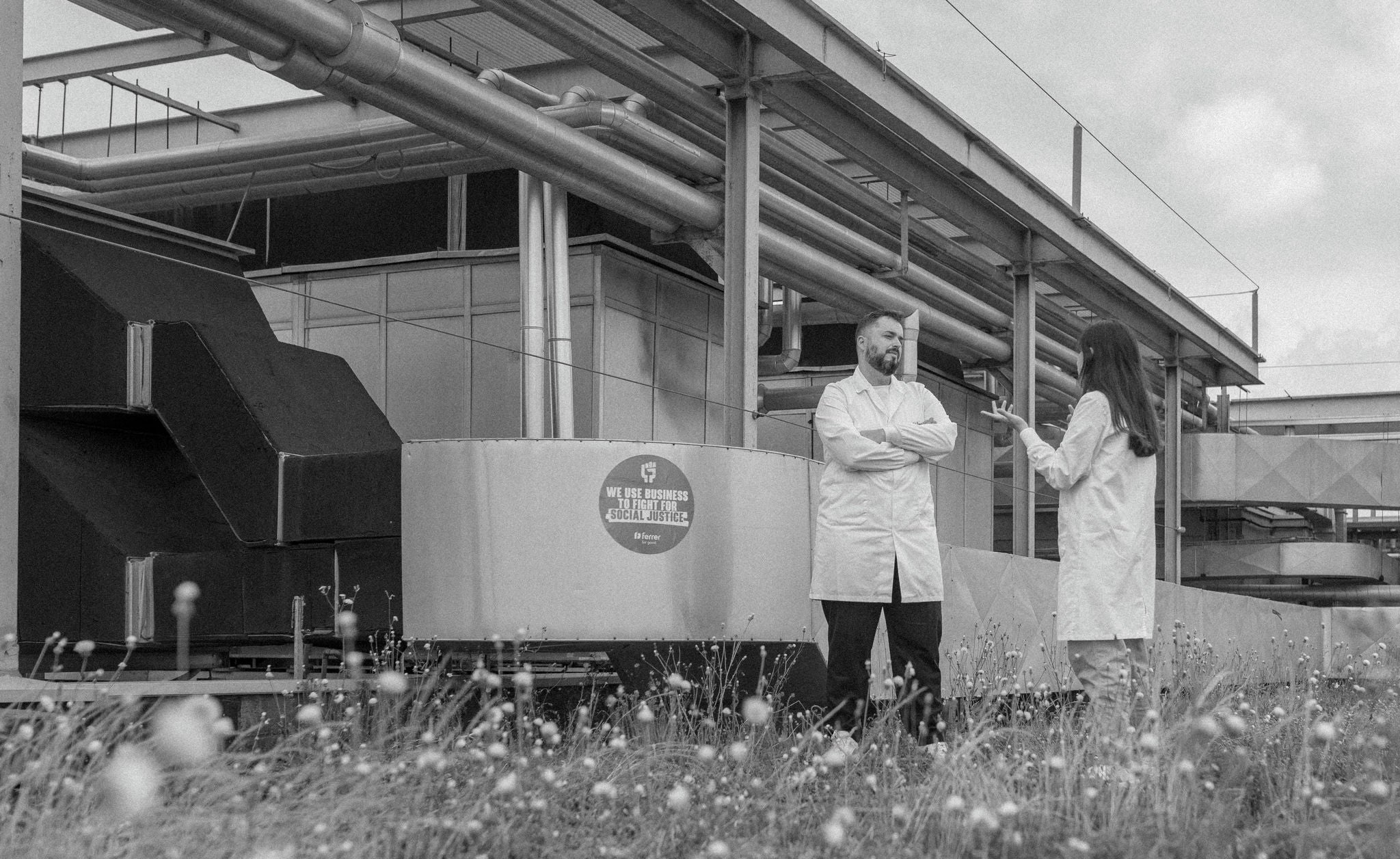 Dos profesionales con bata conversan en una zona industrial exterior, junto a tuberías y equipos, con vegetación en primer plano, en una imagen en blanco y negro.