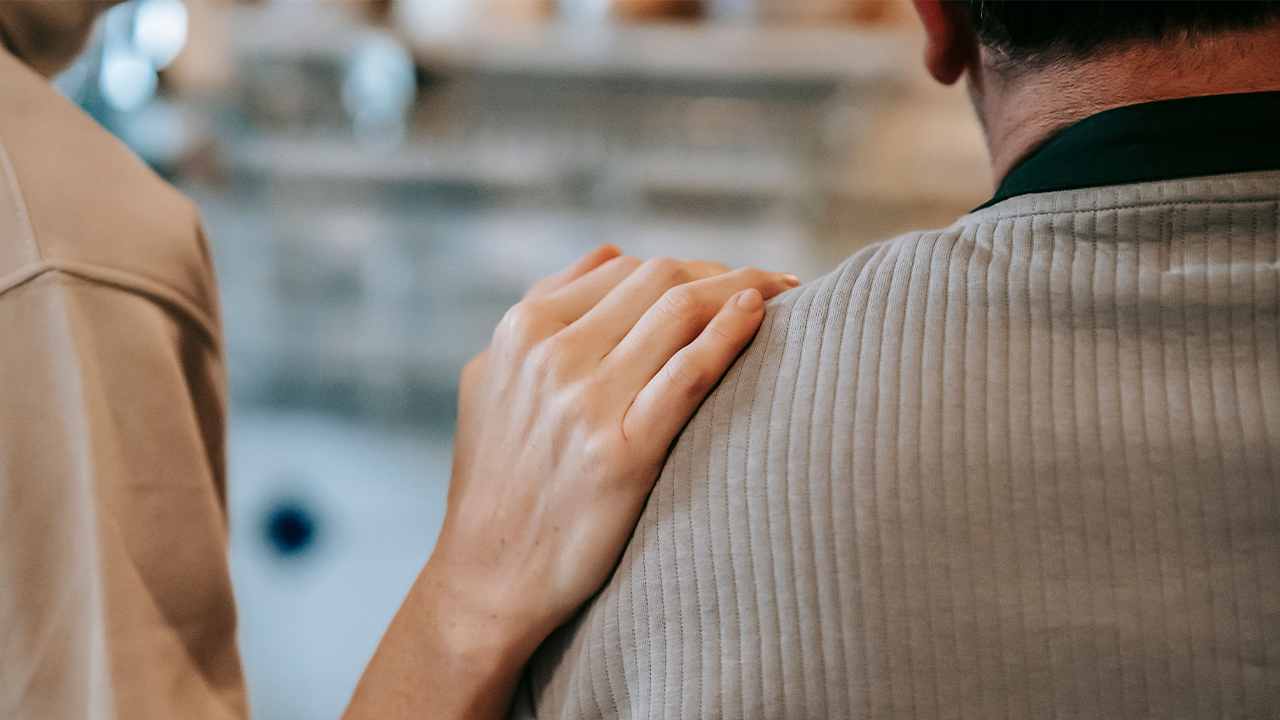 Vista trasera de una persona que coloca suavemente una mano sobre el hombro de otra vestida con un suéter gris, en un gesto de consuelo o apoyo.
