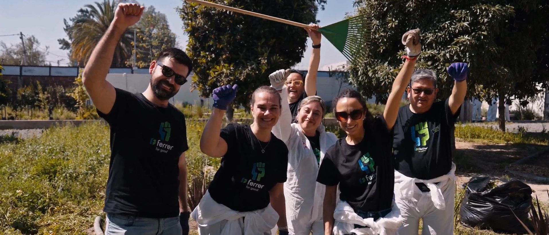 Voluntarios de Ferrer sonríen y saludan a cámara durante una actividad de limpieza al aire libre, equipados con guantes y bolsas de recogida.